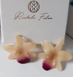 Coppia di orecchini artigianali realizzati con vere orchidee color panna e bianco con un intenso labello sfumato in viola scuro sul bordo inferiore, inglobate in resina lucida e mostrate davanti alla loro scatola regalo bianca con logo Rachele Falvo.