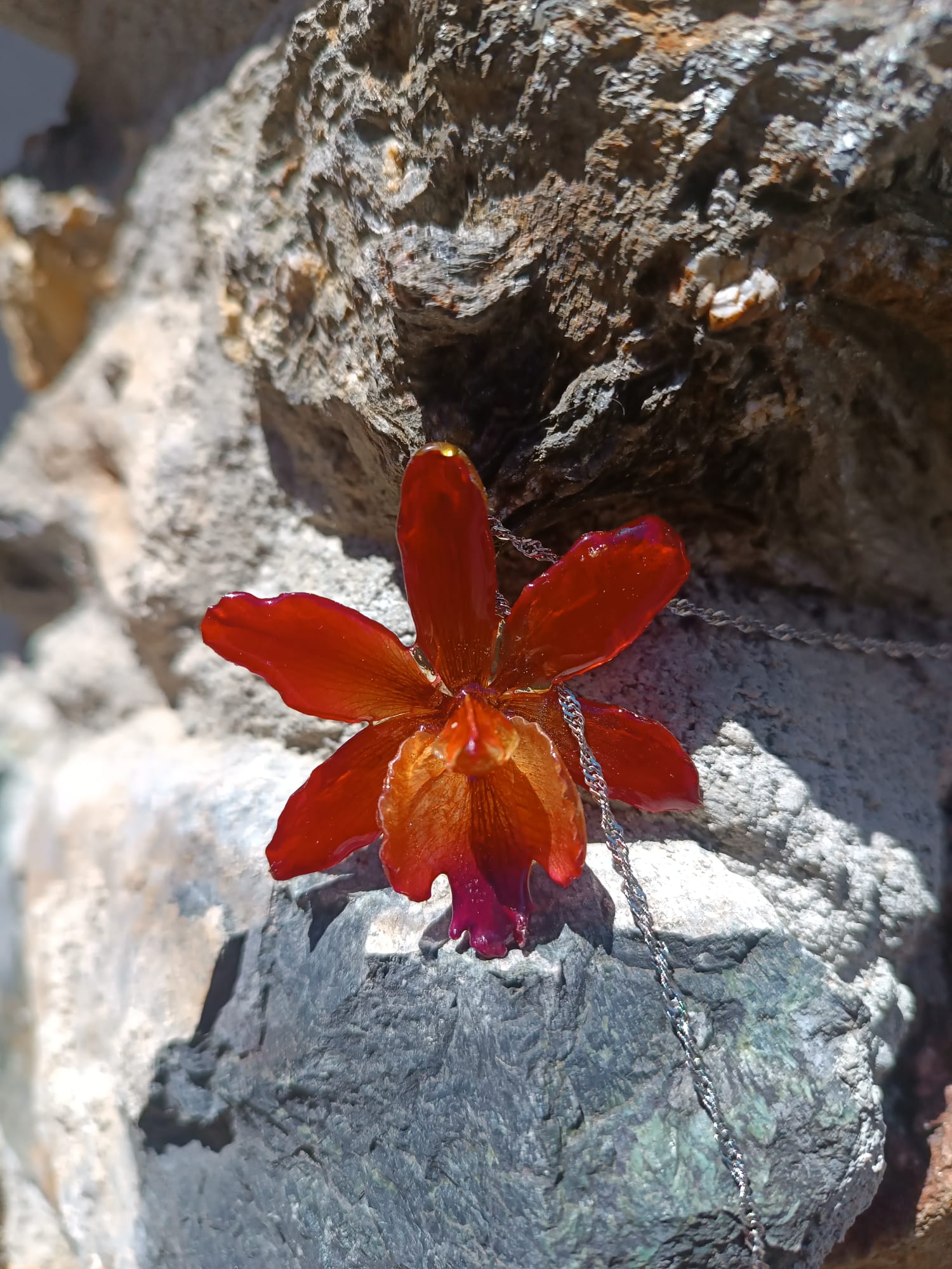 Ciondolo artigianale con vera orchidea marrone e arancio racchiusa in resina trasparente con riflessi di luce, mostrato con catenina in acciaio color oro e color argento.