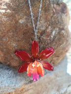 Ciondolo artigianale con vera orchidea marrone e arancio racchiusa in resina trasparente con riflessi di luce, mostrato con catenina in acciaio color oro e color argento.