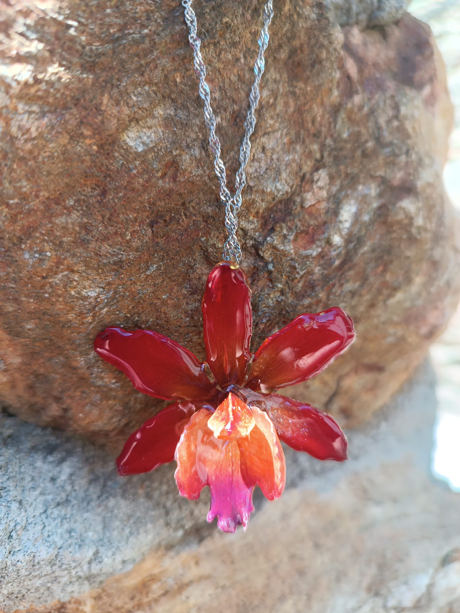 Ciondolo artigianale con vera orchidea marrone e arancio racchiusa in resina trasparente con riflessi di luce, mostrato con catenina in acciaio color oro e color argento.