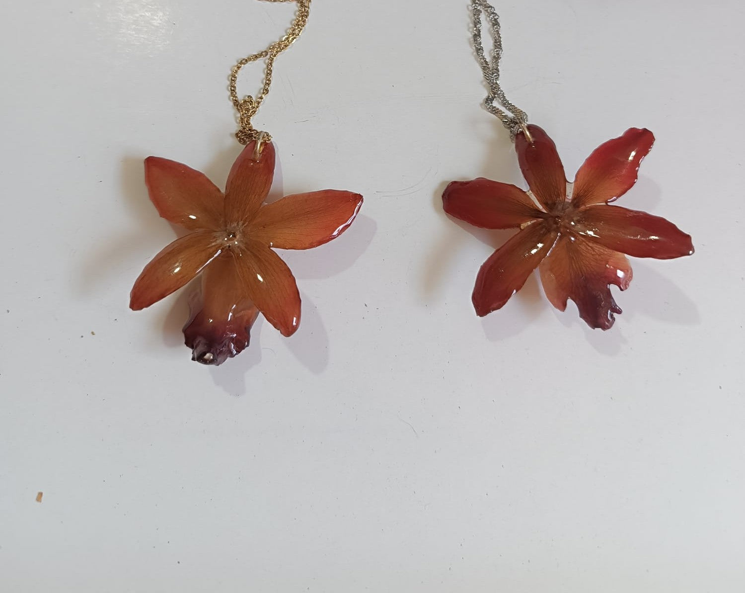 parte posteriore Ciondolo artigianale con vera orchidea marrone e arancio racchiusa in resina trasparente con riflessi di luce, mostrato con catenina in acciaio color oro e color argento.
