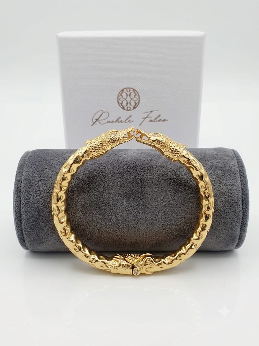 bracciale rigido a molla in bronzo dorato di Rachele Falvo con due teste di serpente e occhi in topazio bianco naturale, poggiato sulla scatola regalo