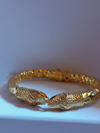 bracciale rigido a molla in bronzo dorato di Rachele Falvo con due teste di serpente e occhi in topazio bianco naturale, poggiato sulla scatola regalo
