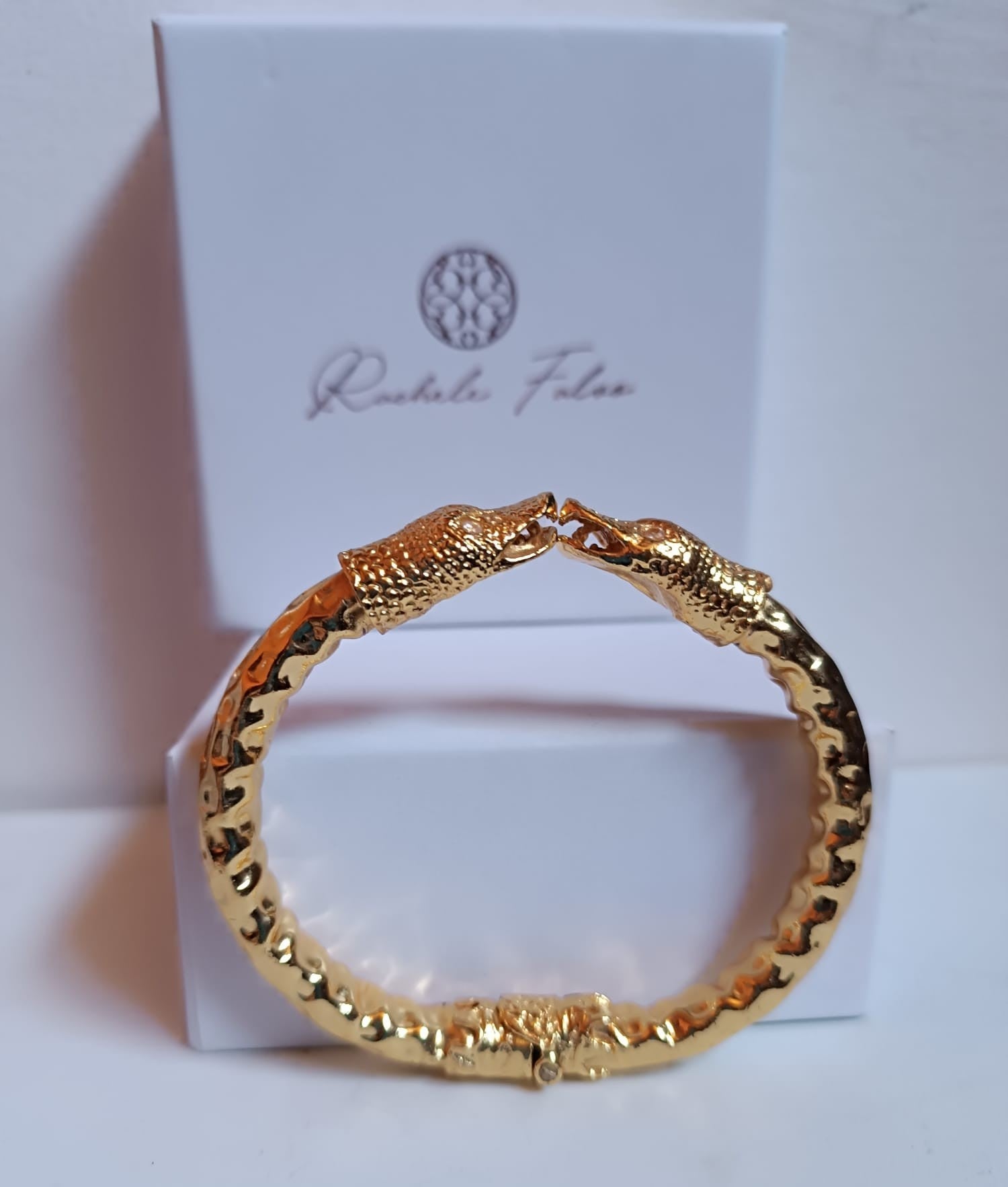 bracciale rigido a molla in bronzo dorato di Rachele Falvo con due teste di serpente e occhi in topazio bianco naturale, poggiato sulla scatola regalo