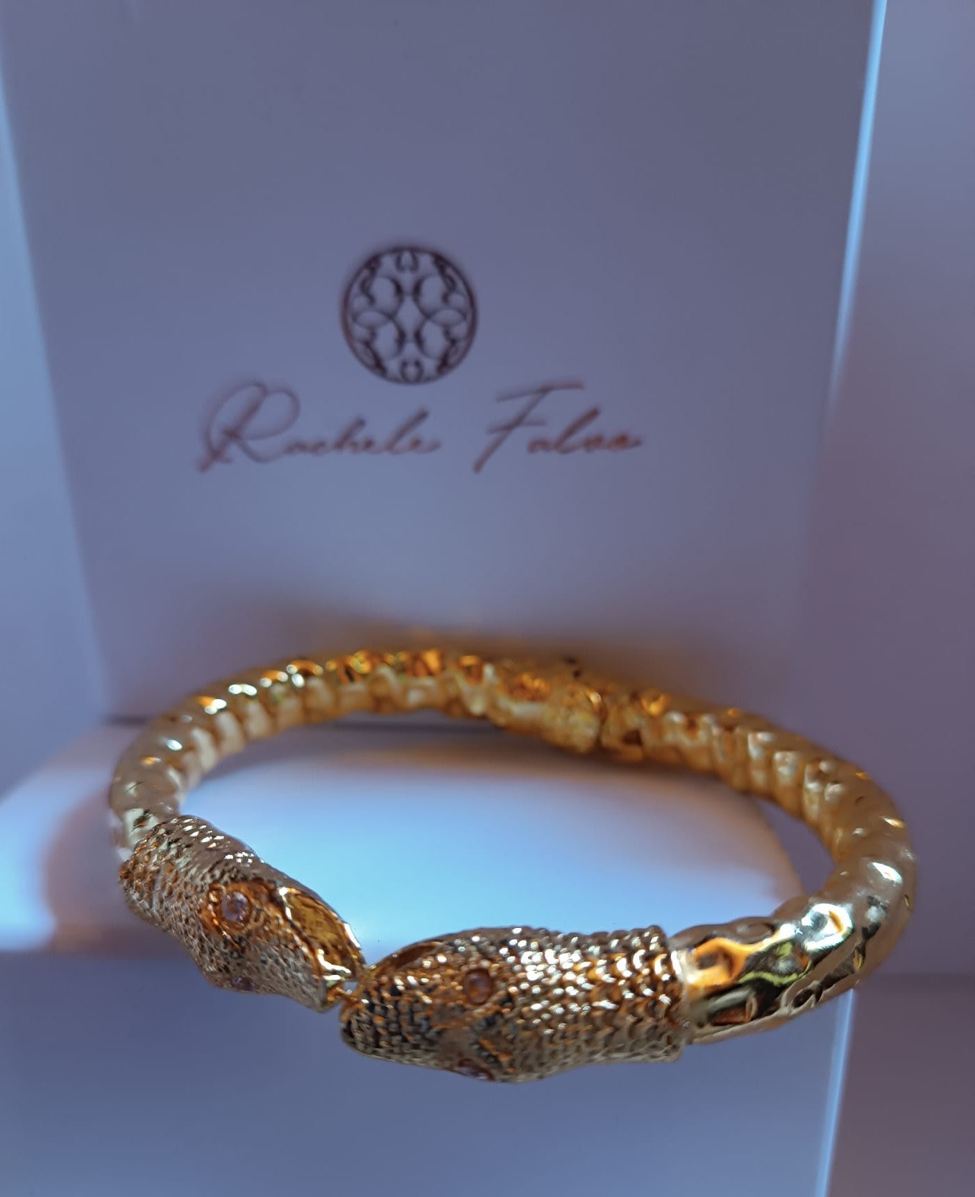 bracciale rigido a molla in bronzo dorato di Rachele Falvo con due teste di serpente e occhi in topazio bianco naturale, poggiato sulla scatola regalo
