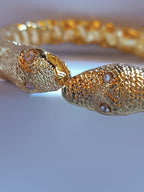 bracciale rigido a molla in bronzo dorato di Rachele Falvo con due teste di serpente e occhi in topazio bianco naturale, poggiato sulla scatola regalo