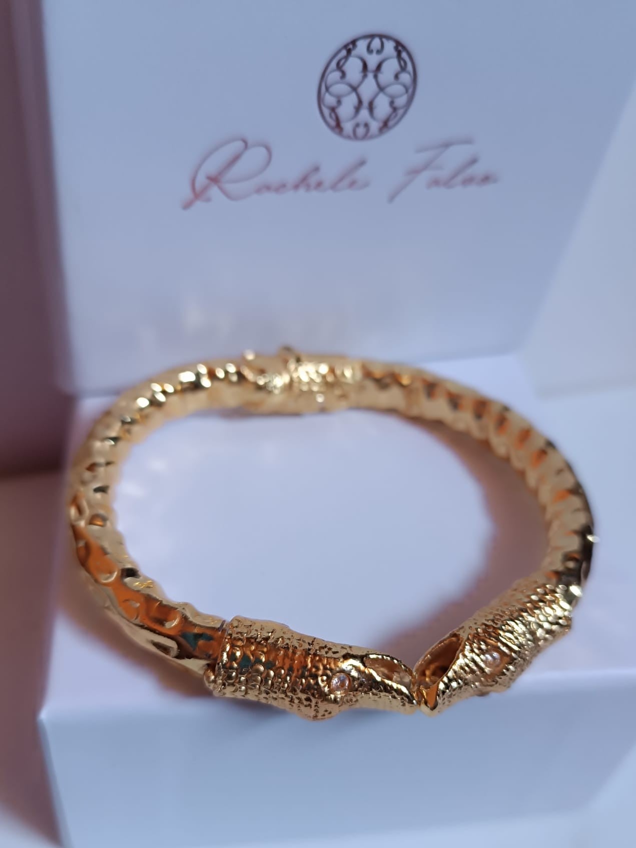bracciale rigido a molla in bronzo dorato di Rachele Falvo con due teste di serpente e occhi in topazio bianco naturale, poggiato sulla scatola regalo