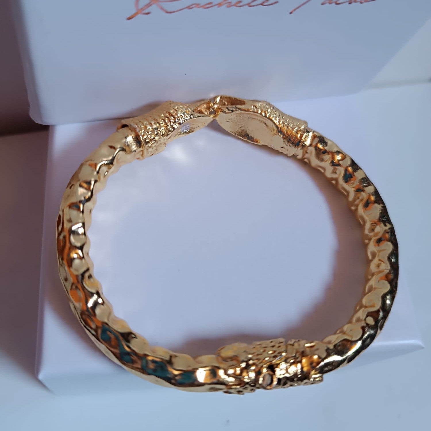 bracciale rigido a molla in bronzo dorato di Rachele Falvo con due teste di serpente e occhi in topazio bianco naturale, poggiato sulla scatola regalo