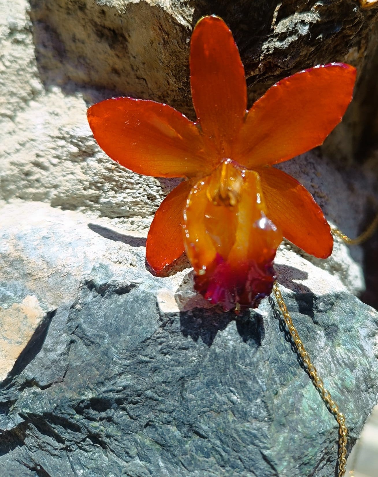 Ciondolo artigianale con vera orchidea marrone e arancio racchiusa in resina trasparente con riflessi di luce, mostrato con catenina in acciaio color oro e color argento.
