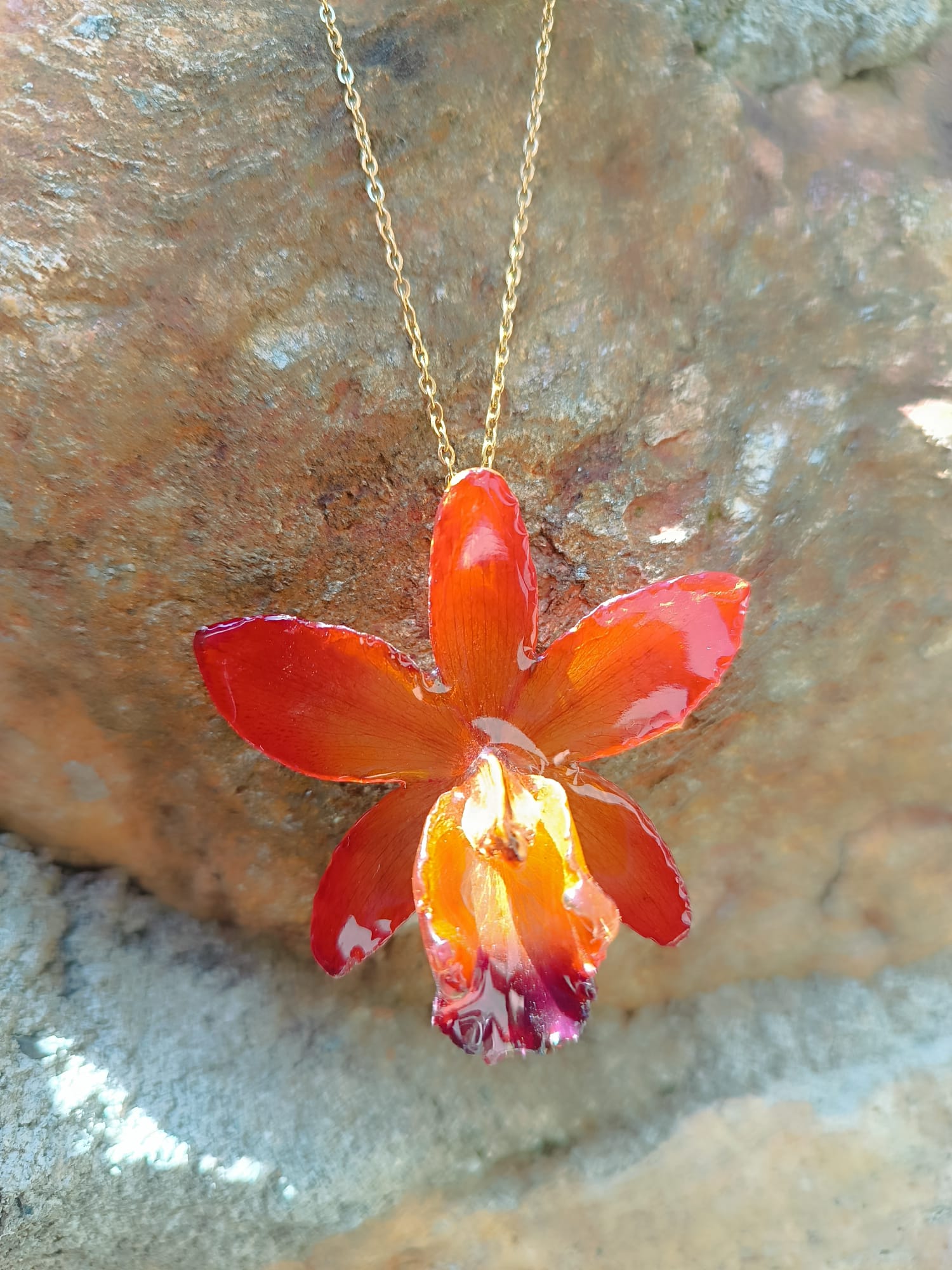 Ciondolo artigianale con vera orchidea marrone e arancio racchiusa in resina trasparente con riflessi di luce, mostrato con catenina in acciaio color oro e color argento.