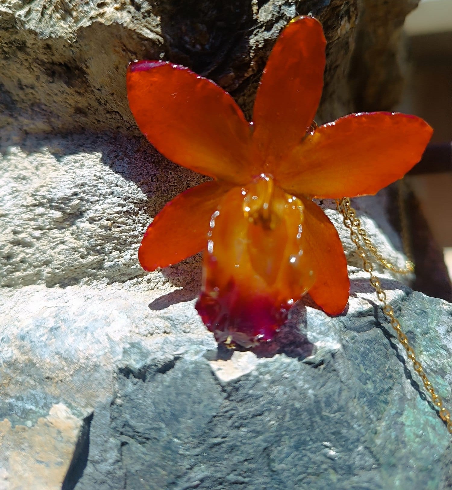 Ciondolo artigianale con vera orchidea marrone e arancio racchiusa in resina trasparente con riflessi di luce, mostrato con catenina in acciaio color oro e color argento.