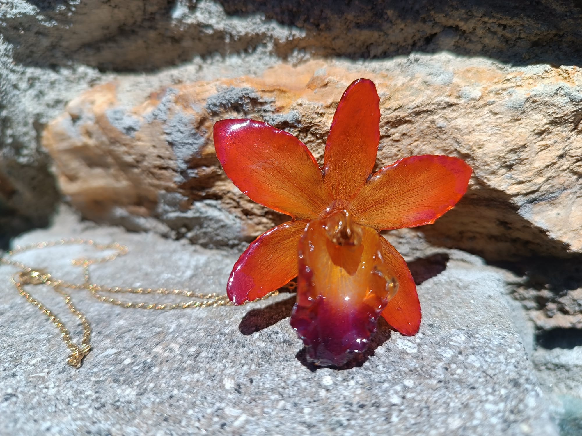 Ciondolo artigianale con vera orchidea marrone e arancio racchiusa in resina trasparente con riflessi di luce, mostrato con catenina in acciaio color oro e color argento.