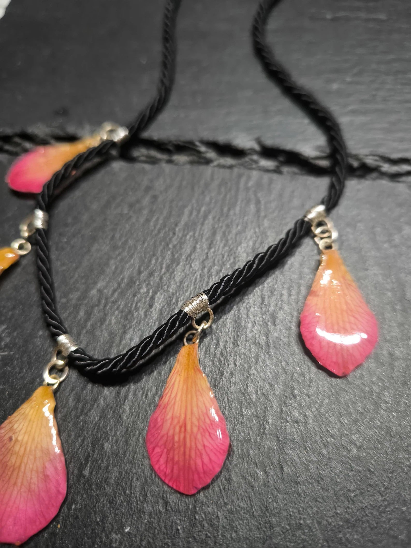 Primo piano di una collana artigianale in seta intrecciata color charcoal con cinque pendenti a goccia in resina trasparente contenenti petali di vera orchidea rosa e crema