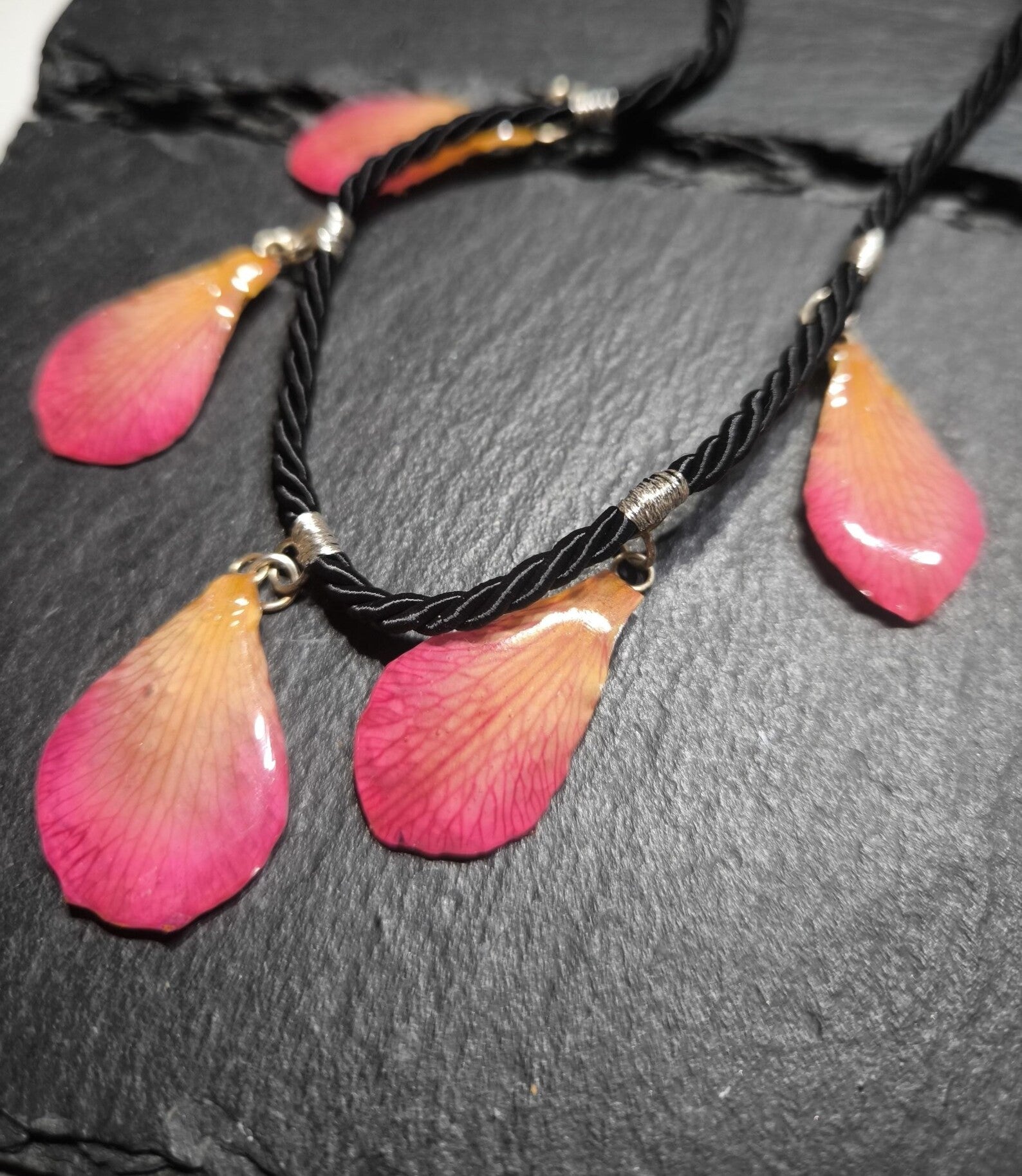 Primo piano di una collana artigianale in seta intrecciata color charcoal con cinque pendenti a goccia in resina trasparente contenenti petali di vera orchidea rosa e crema