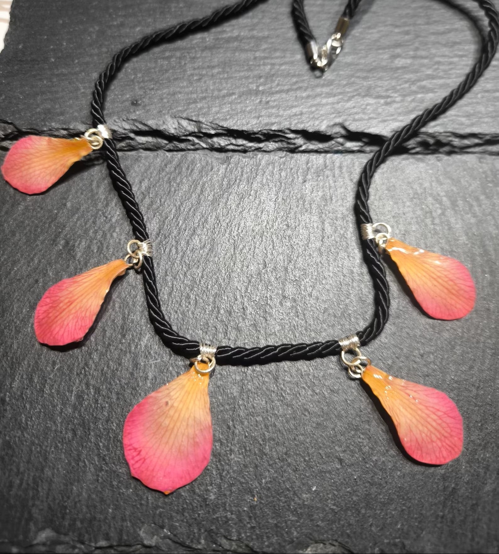 Primo piano di una collana artigianale in seta intrecciata color charcoal con cinque pendenti a goccia in resina trasparente contenenti petali di vera orchidea rosa e crema