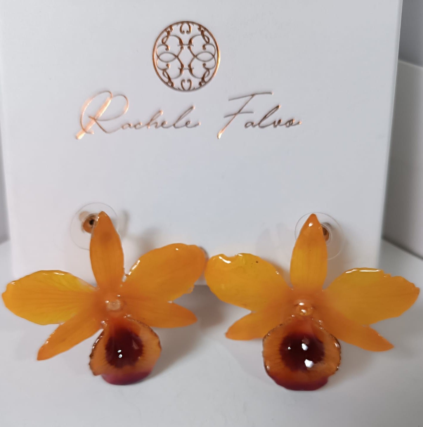 orecchini artigianali al lobo realizzati con intere orchidee vere in resina lucida, una variante  giallo ocra con sfumature viola, disposte sulla loro scatola regalo bianca firmata Rachele Falvo.