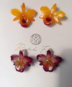 orecchini artigianali al lobo realizzati con intere orchidee vere in resina lucida, una variante viola ed una variante  giallo ocra con sfumature viola, disposte sulla loro scatola regalo bianca firmata Rachele Falvo.