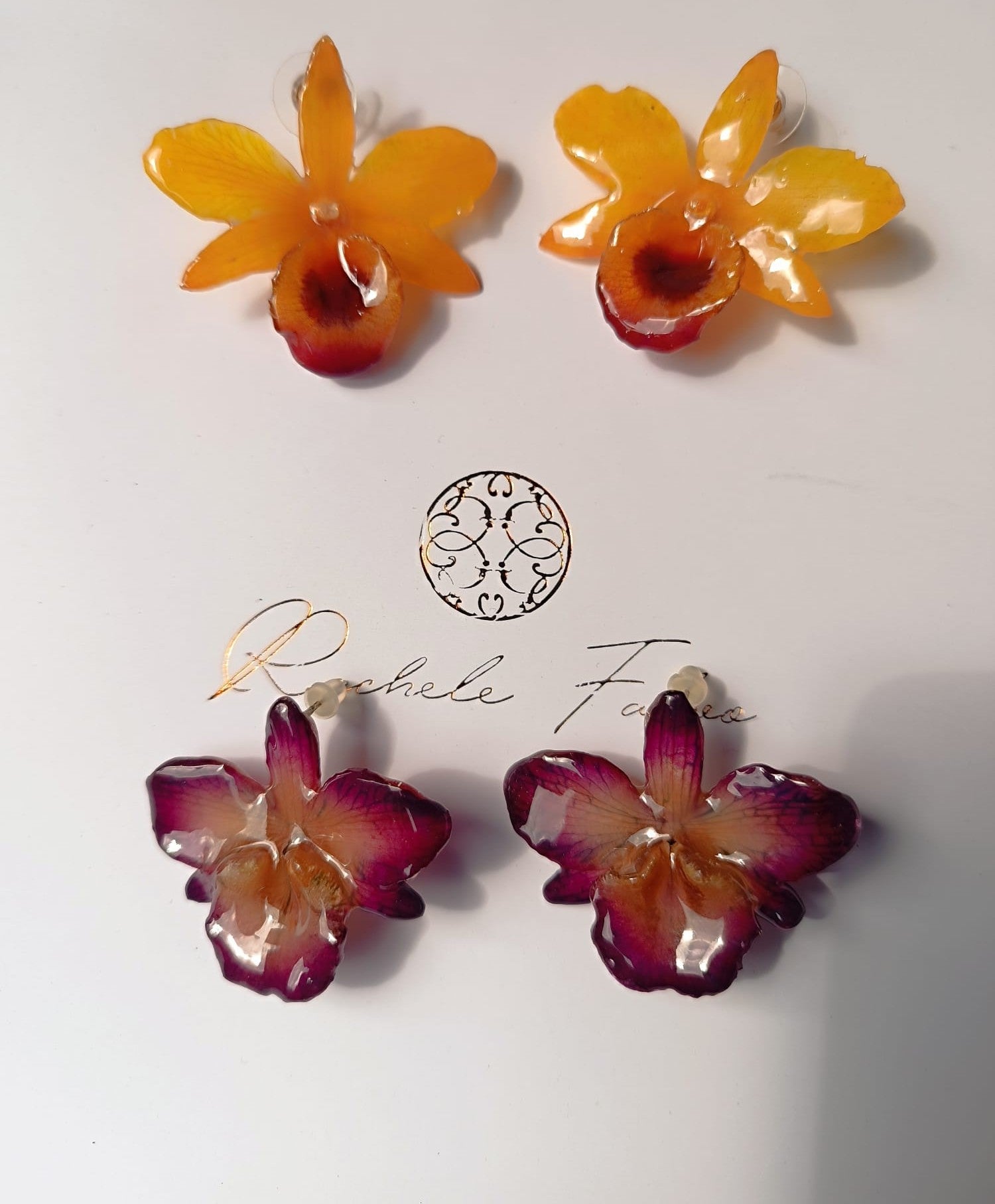 orecchini artigianali al lobo realizzati con intere orchidee vere in resina lucida, una variante viola ed una variante  giallo ocra con sfumature viola, disposte sulla loro scatola regalo bianca firmata Rachele Falvo.