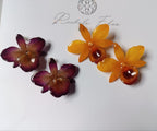 Due coppie di grandi orecchini artigianali al lobo realizzati con intere orchidee vere in resina lucida, una variante viola scuro e una variante giallo ocra con sfumature marroni, disposte sulla loro scatola regalo bianca firmata Rachele Falvo.
