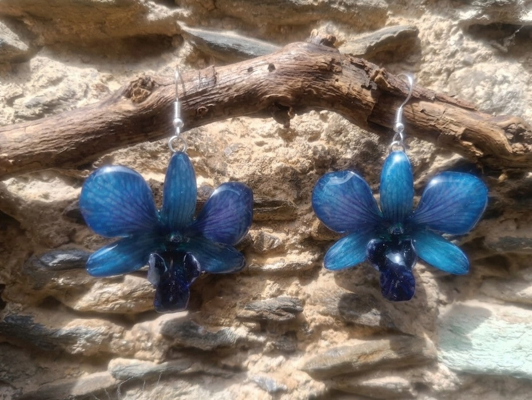 Orecchini pendenti con vere orchidee blu e venature lilla inglobate in resina trasparente, fotografati su sfondo neutro per mostrare la trasparenza e i dettagli del fiore.