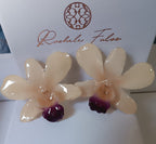 Coppia di orecchini pendenti realizzati artigianalmente con vere orchidee bianche Dendrobium racchiuse in resina trasparente lucida con ganci in argento, presentati accanto alla loro scatola regalo.