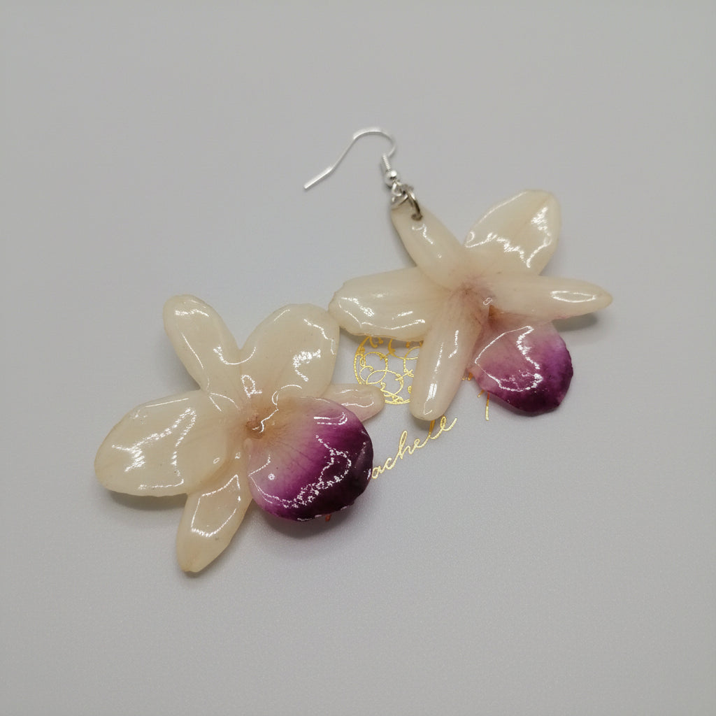 Coppia di orecchini artigianali realizzati con vere orchidee color panna e bianco con un intenso labello sfumato in viola scuro sul bordo inferiore, inglobate in resina lucida e mostrate davanti alla loro scatola regalo bianca con logo Rachele Falvo.