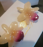Coppia di orecchini artigianali realizzati con vere orchidee color panna e bianco con un intenso labello sfumato in viola scuro sul bordo inferiore, inglobate in resina lucida e mostrate davanti alla loro scatola regalo bianca con logo Rachele Falvo.