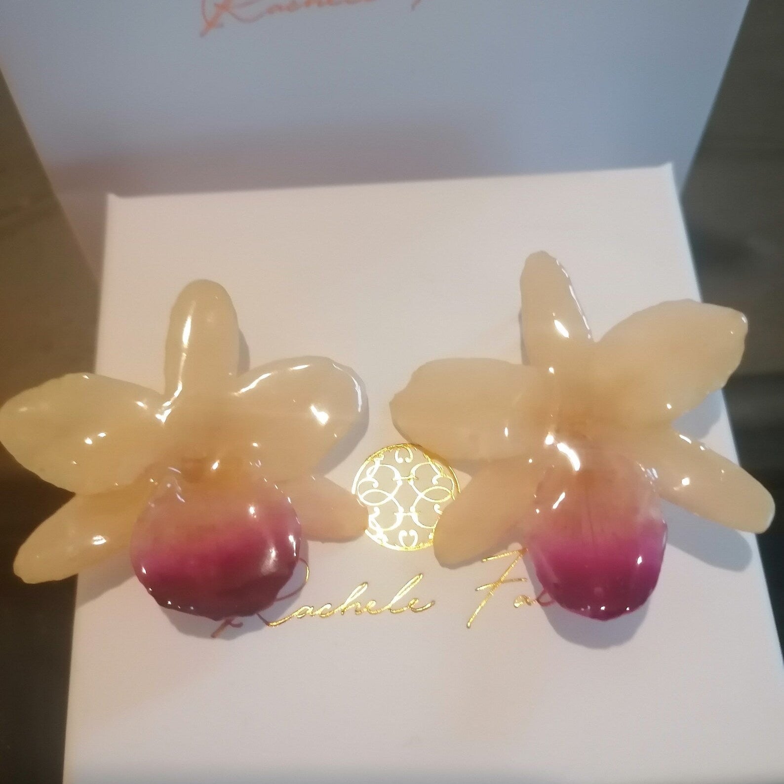 Coppia di orecchini artigianali realizzati con vere orchidee color panna e bianco con un intenso labello sfumato in viola scuro sul bordo inferiore, inglobate in resina lucida e mostrate davanti alla loro scatola regalo bianca con logo Rachele Falvo.