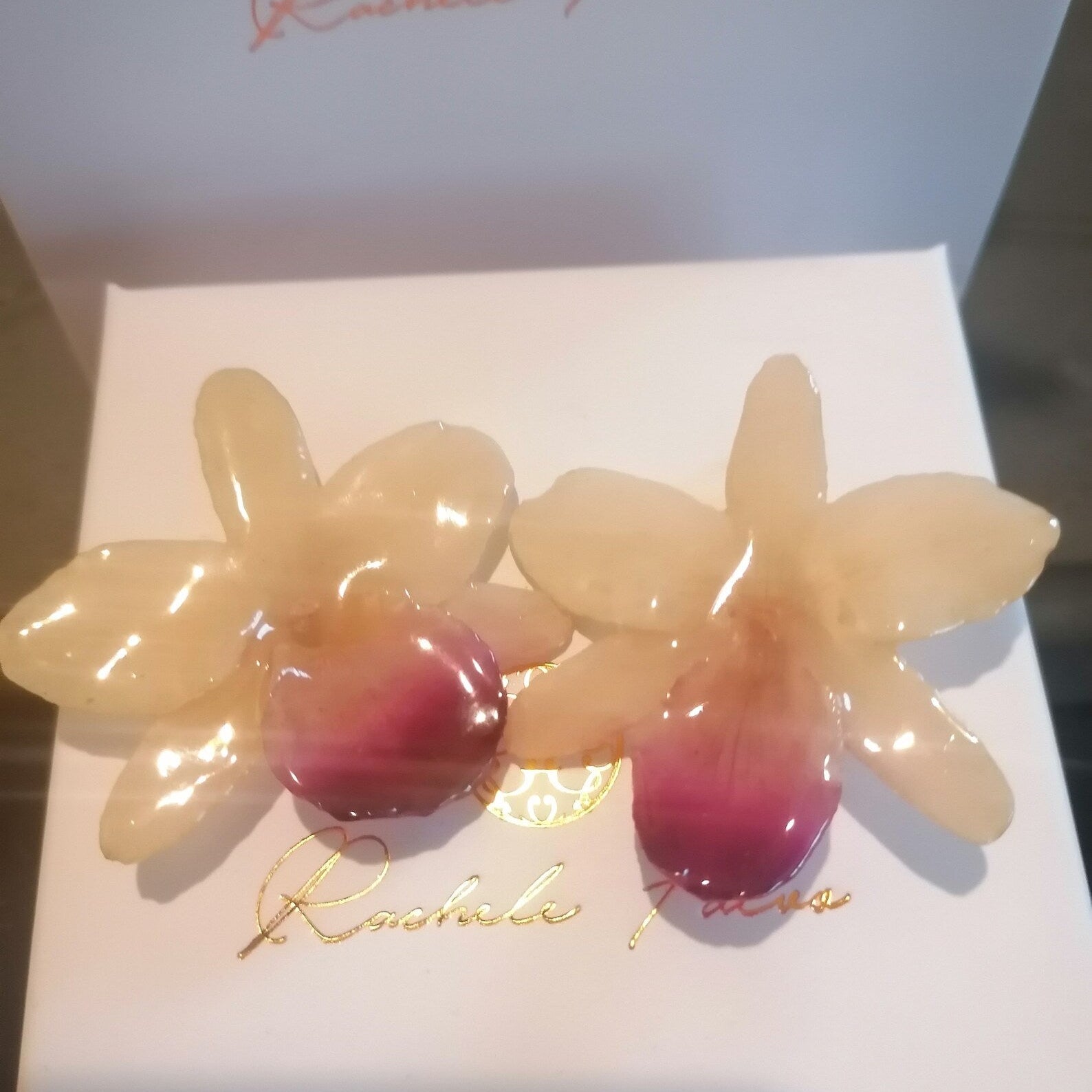 Coppia di orecchini artigianali realizzati con vere orchidee color panna e bianco con un intenso labello sfumato in viola scuro sul bordo inferiore, inglobate in resina lucida e mostrate davanti alla loro scatola regalo bianca con logo Rachele Falvo.