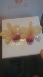 Coppia di orecchini artigianali realizzati con vere orchidee color panna e bianco con un intenso labello sfumato in viola scuro sul bordo inferiore, inglobate in resina lucida e mostrate davanti alla loro scatola regalo bianca con logo Rachele Falvo.
