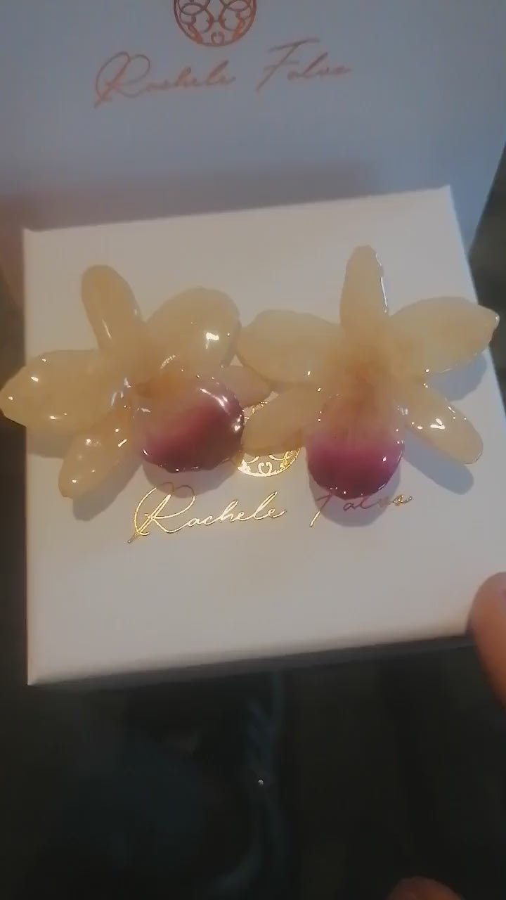 Coppia di orecchini artigianali realizzati con vere orchidee color panna e bianco con un intenso labello sfumato in viola scuro sul bordo inferiore, inglobate in resina lucida e mostrate davanti alla loro scatola regalo bianca con logo Rachele Falvo.
